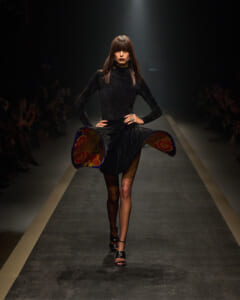Model walking the runway in a black velvet dress with colorful patterned lining peeking from the sides under a spotlight on a dark catwalk.