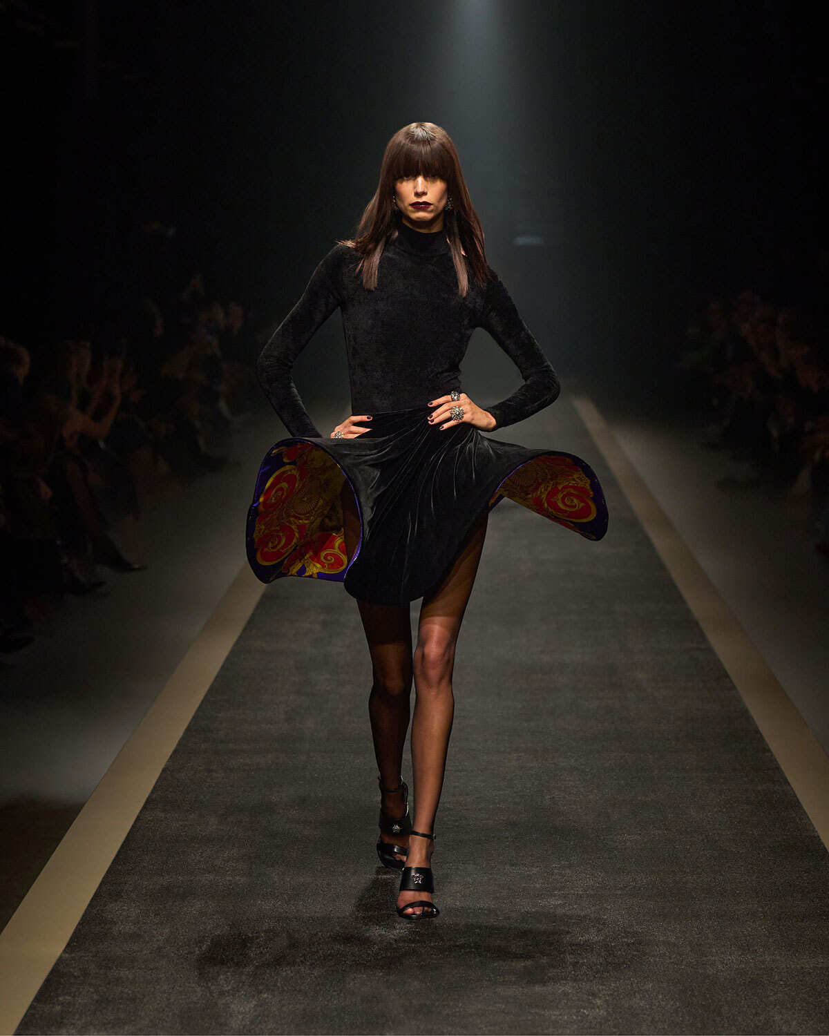 Model walking the runway in a black velvet dress with colorful patterned lining peeking from the sides under a spotlight on a dark catwalk.