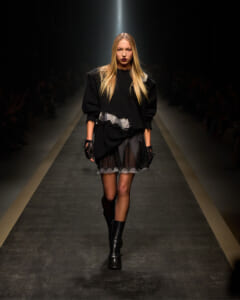Model walking a fashion show runway in an all-black outfit: oversized sweater, lace-trimmed skirt, gloves, and knee-high boots under spotlight.