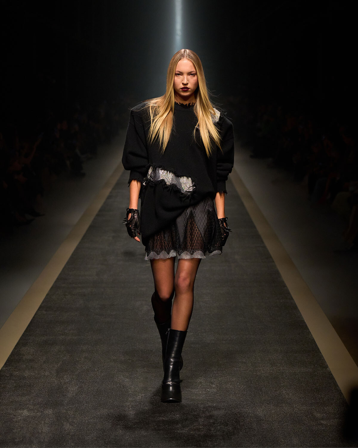 Model walking a fashion show runway in an all-black outfit: oversized sweater, lace-trimmed skirt, gloves, and knee-high boots under spotlight.