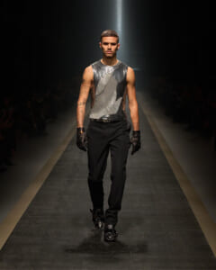 Male model walks the runway in a metallic silver sleeveless top, black pants, and black gloves; spotlight on him with a dark audience backdrop.