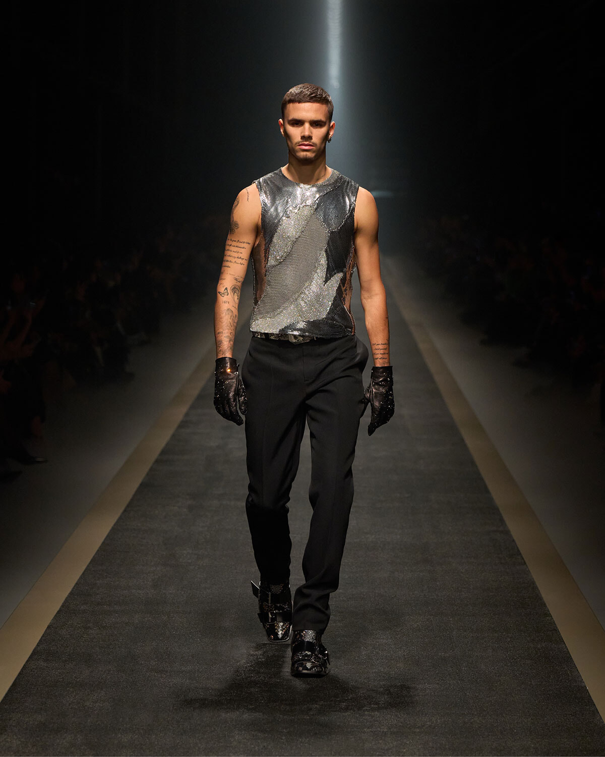 Male model walks the runway in a metallic silver sleeveless top, black pants, and black gloves; spotlight on him with a dark audience backdrop.