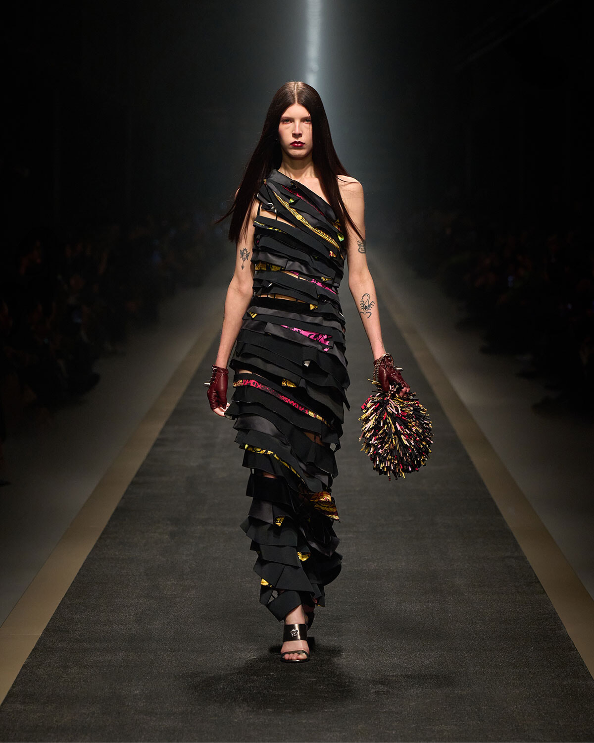 Model walks the runway in a black, layered dress with colorful ribbons, carrying a spiky multicolor pom-pom clutch.