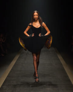 Model walking a runway in a black off-shoulder dress, lifting the skirt to reveal a gold patterned lining on the inside.