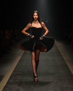 Model walks a fashion runway in a strapless black dress with a voluminous skirt and bold jewelry.