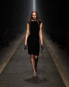 Model walking a runway in a black velvet sheath dress with metallic collar, gloves, and sparkly accessories under a spotlight on a dark catwalk