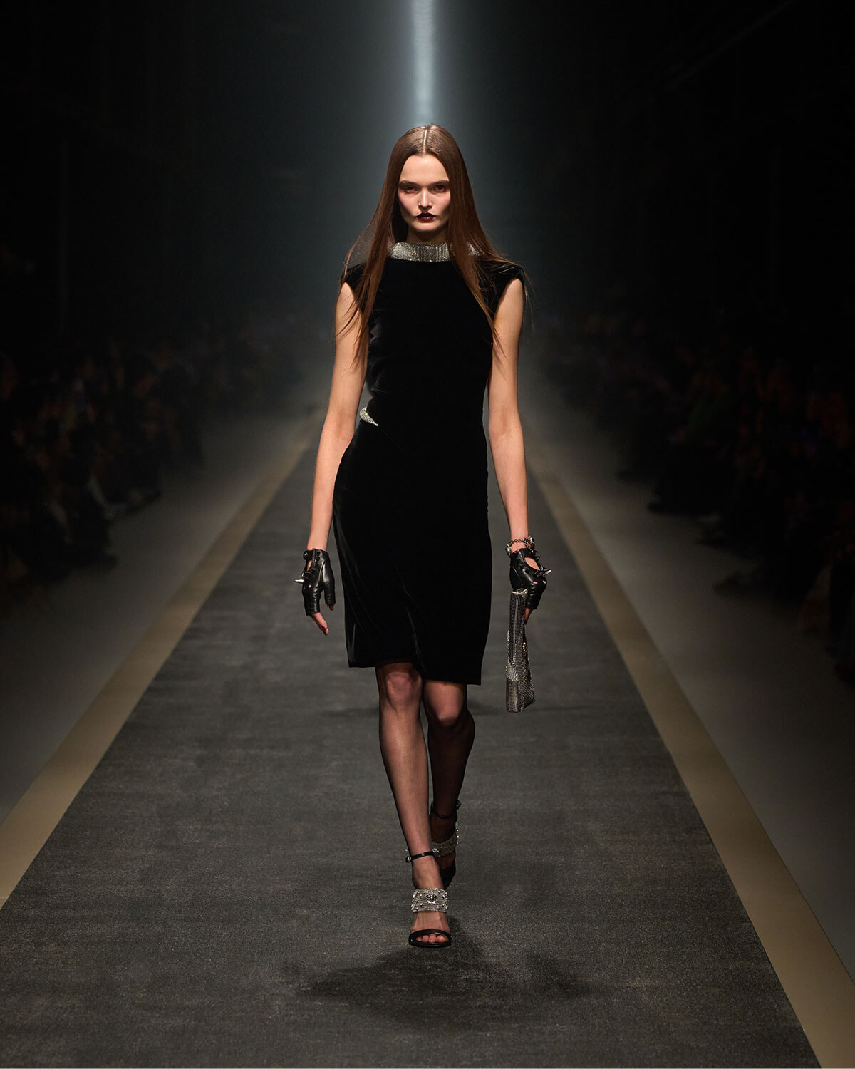 Model walking a runway in a black velvet sheath dress with metallic collar, gloves, and sparkly accessories under a spotlight on a dark catwalk