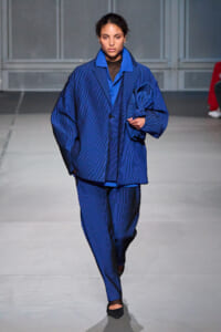 Model walks a runway in an oversized blue pinstripe suit with matching shirt, black tights, and pointed flats; hair pulled back, neutral expression.