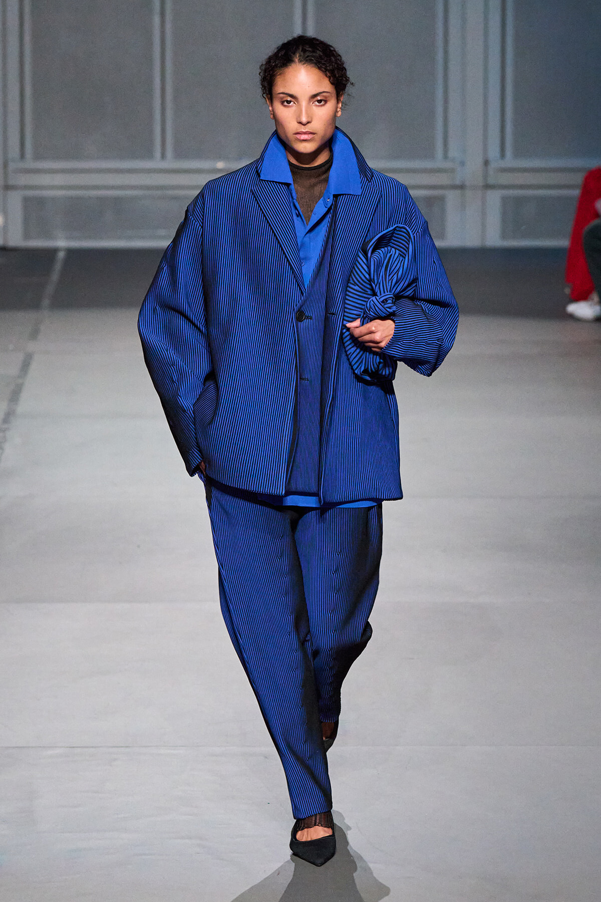 Model walks a runway in an oversized blue pinstripe suit with matching shirt, black tights, and pointed flats; hair pulled back, neutral expression.