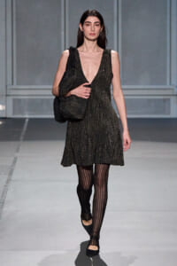 Model walking a runway in a sleeveless, glittery black V-neck dress, carrying a matching black textured bag and wearing striped tights with black flats.