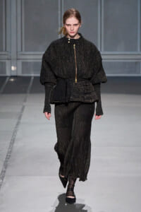 Model on a runway wearing a black glittery suit with a voluminous top and gold zipper detail, paired with wide-leg pants.