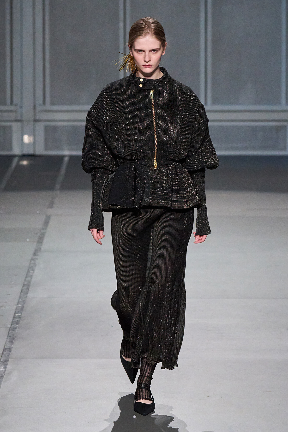 Model on a runway wearing a black glittery suit with a voluminous top and gold zipper detail, paired with wide-leg pants.