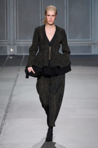 Model on a runway wearing a metallic flecked black peplum suit with a deep V-neck and gold zipper, carrying a black clutch bag