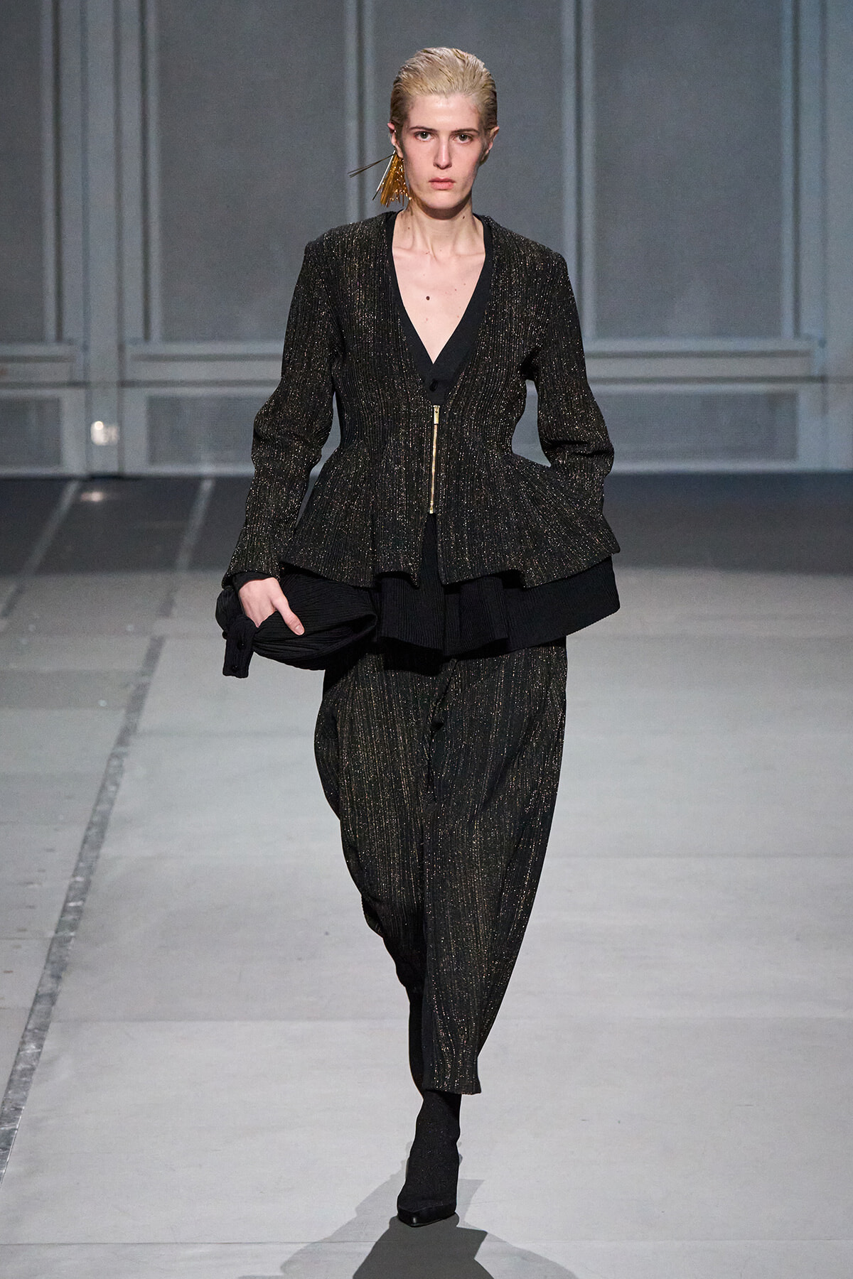 Model on a runway wearing a metallic flecked black peplum suit with a deep V-neck and gold zipper, carrying a black clutch bag