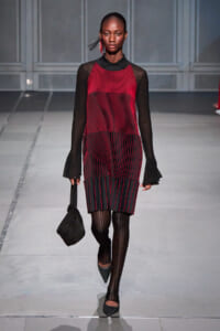 Model on a runway wearing a red-and-black striped dress with sheer black sleeves, black tights, and pointed-toe heels, carrying a black handbag.