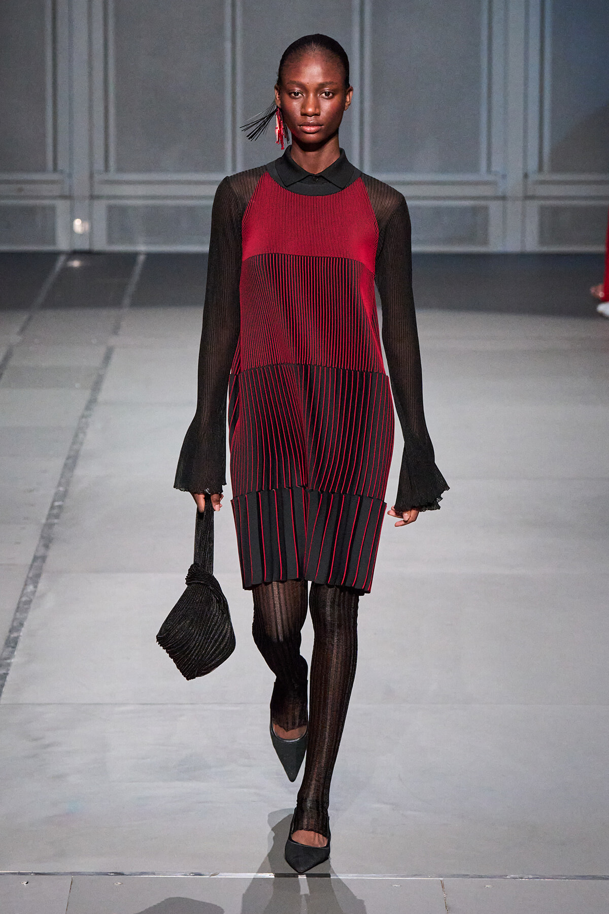 Model on a runway wearing a red-and-black striped dress with sheer black sleeves, black tights, and pointed-toe heels, carrying a black handbag.