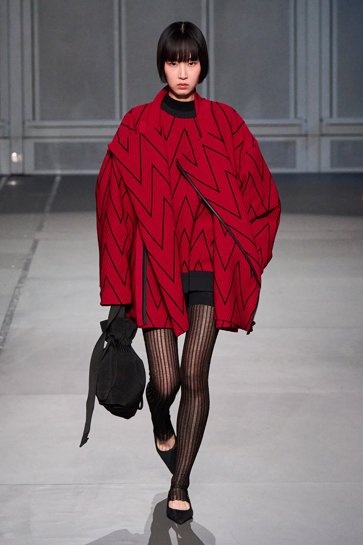 Model walking the runway in an oversized red zigzag-patterned cape over a black outfit, carrying a black drawstring bag.