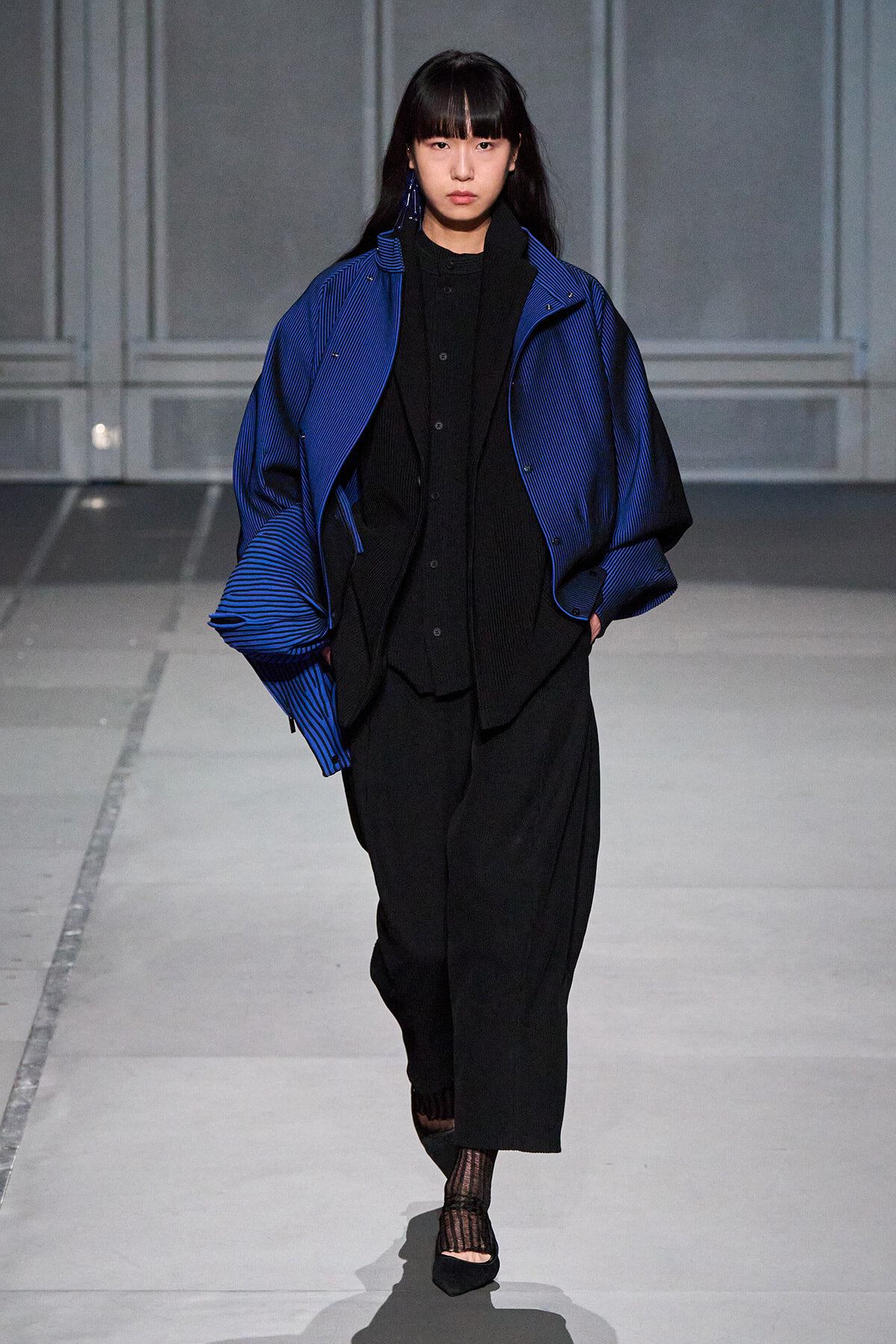 Model walking the runway in an oversized cobalt blue striped jacket over a black layered outfit and wide black pants.