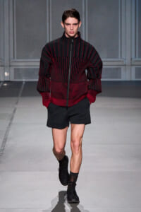 Male model walking a runway in a black and red striped zip-up sweater with oversized sleeves, paired with black shorts and black sneakers.