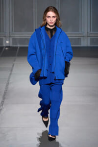 Runway model in a bold cobalt blue oversized jacket with a matching blue ribbed suit and a black sheer top underneath.