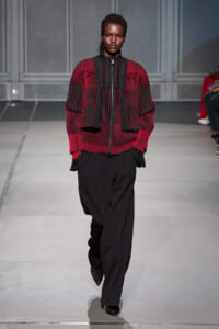 Model walks the runway in a red and black striped knit top with a matching cropped vest and wide black trousers.