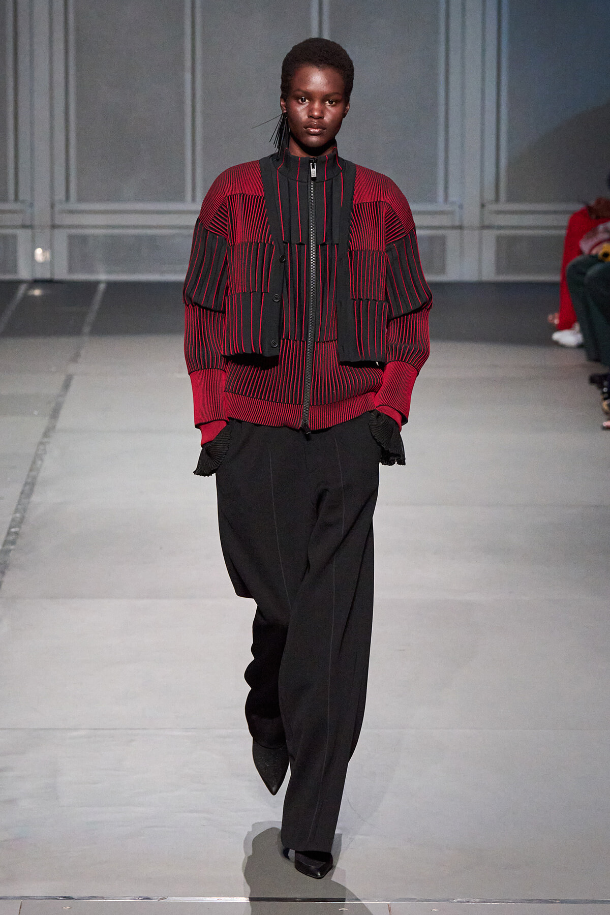 Model walks the runway in a red and black striped knit top with a matching cropped vest and wide black trousers.