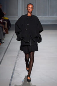 Model walking a runway in an oversized black cardigan, black skirt, and striped tights with black flats, glasses on.