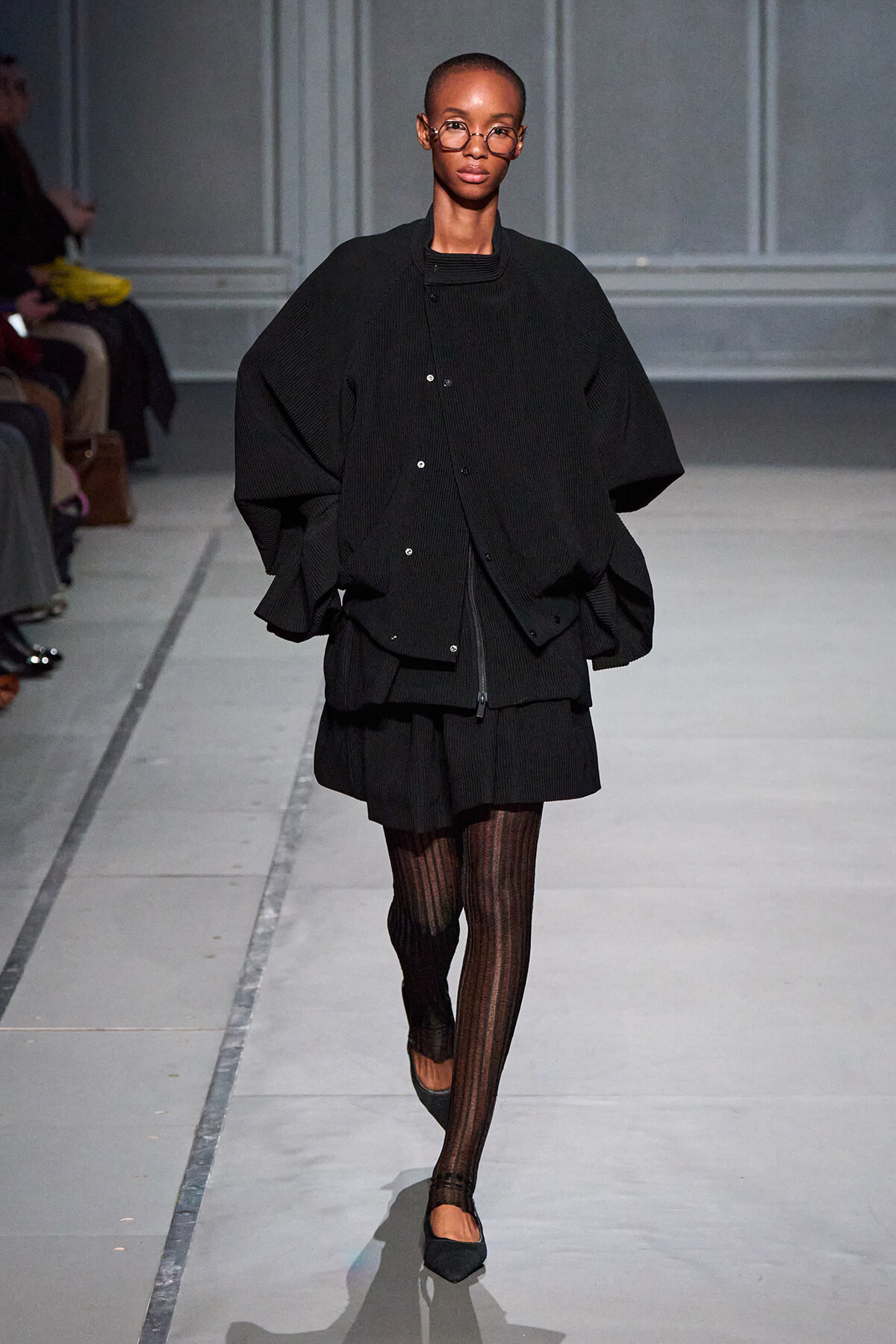 Model walking a runway in an oversized black cardigan, black skirt, and striped tights with black flats, glasses on.