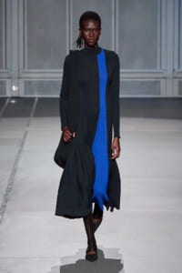 Model walking a runway in a long black coat with a bright blue vertical panel down the front.