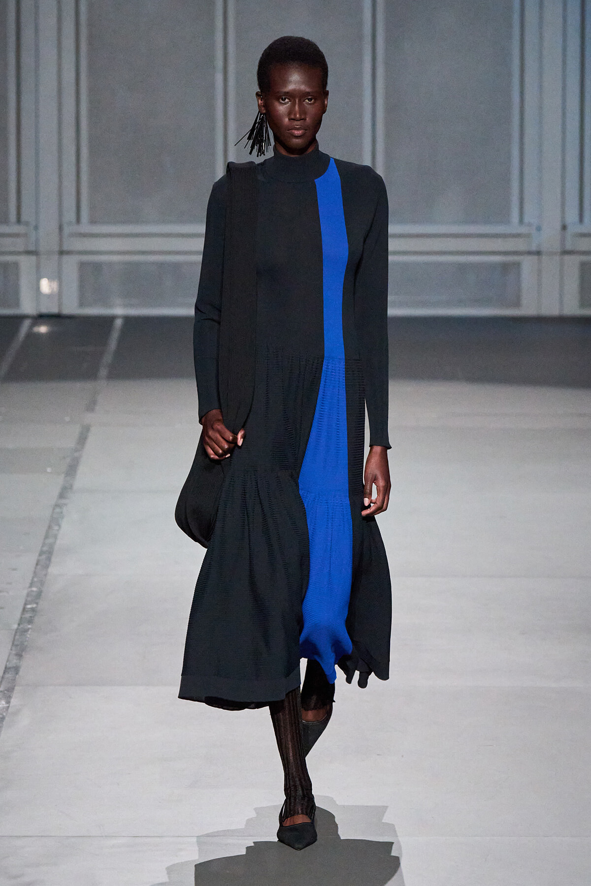 Model walking a runway in a long black coat with a bright blue vertical panel down the front.