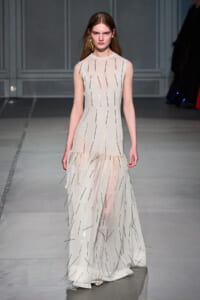 Model on a runway wearing a sleeveless sheer cream gown adorned with slender gold and dark metal rods.