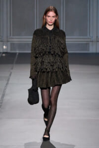 Model walking a runway in a sparkly black layered dress with fringe detailing, striped tights, and black flats, carrying a matching knit bag.