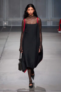 Model walking a runway in a black slip dress layered over a sheer striped turtleneck, red straps at the shoulders, and a small black bag.