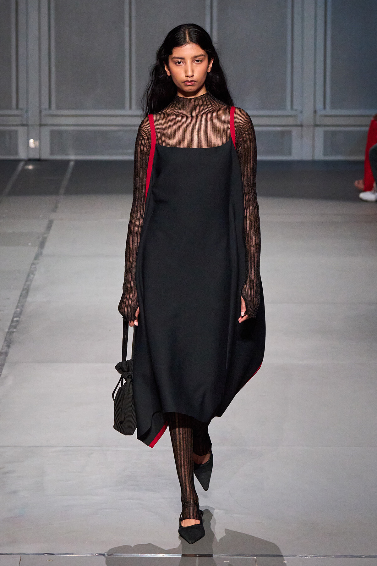 Model walking a runway in a black slip dress layered over a sheer striped turtleneck, red straps at the shoulders, and a small black bag.