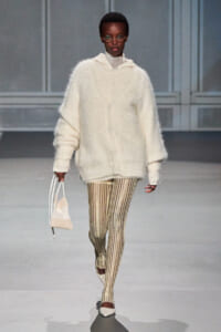 Model on a runway in an oversized white fuzzy cardigan, gold striped tights, cream flats, round glasses, and a small beige handbag.