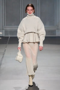 Model on a runway wearing a cream knit turtleneck with a layered peplum and a sheer knit skirt, carrying a small cream bag.