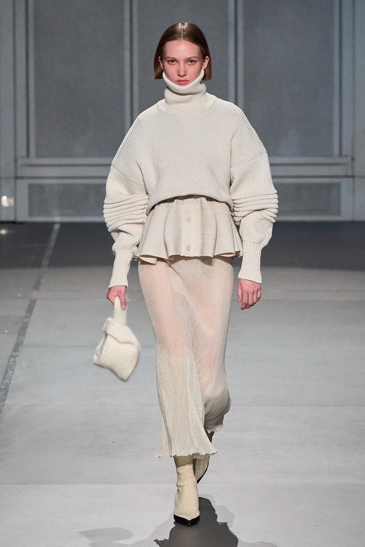 Model on a runway wearing a cream knit turtleneck with a layered peplum and a sheer knit skirt, carrying a small cream bag.