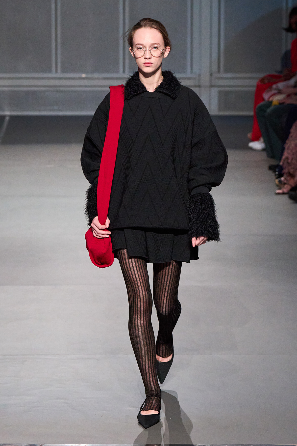 Model walks runway in oversized black knit top with zigzag stitching, black fluffy cuffs, matching skirt, striped tights, black flats, and a red crossbody bag.