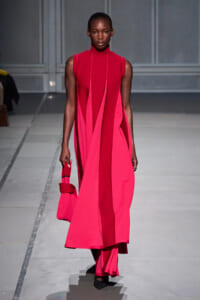 Model on a runway wearing a sleeveless bright pink color-block dress with vertical panels, carrying a small pink handbag.