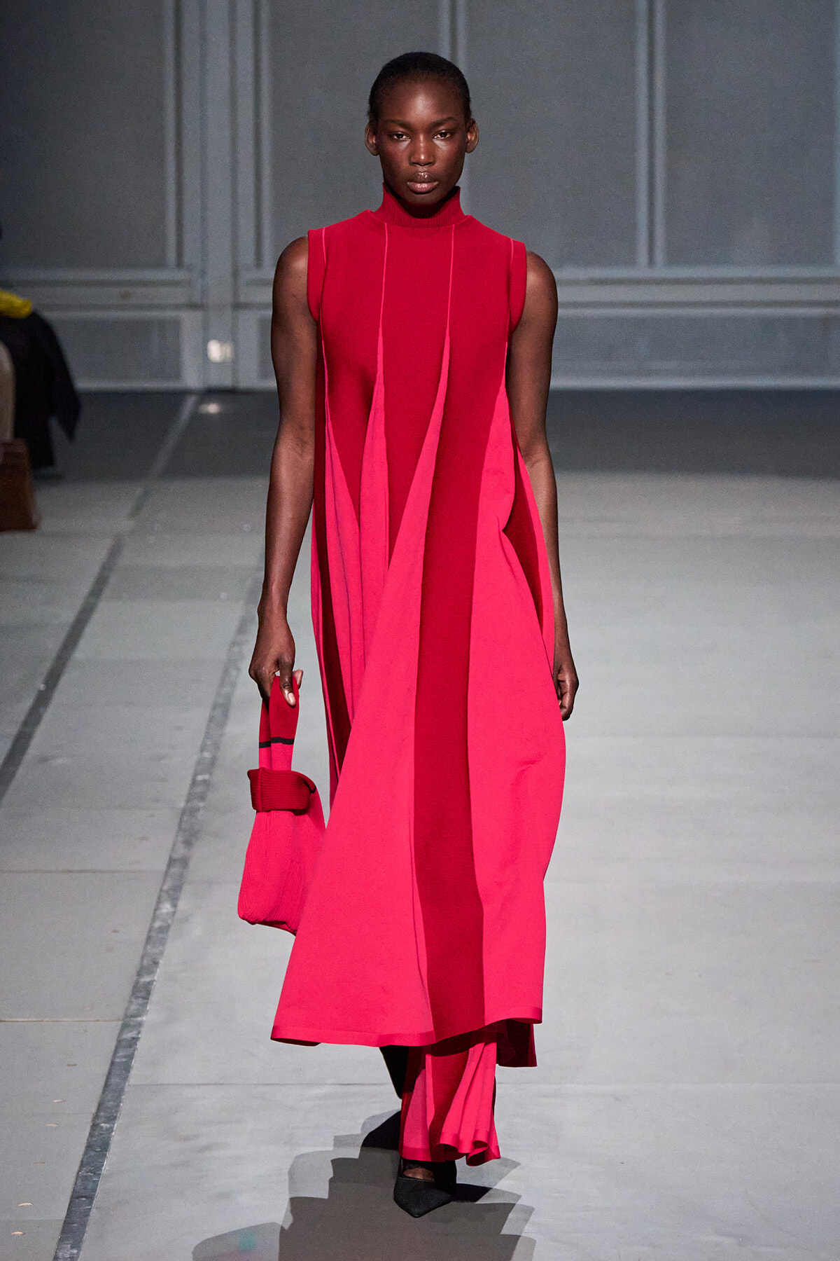 Model on a runway wearing a sleeveless bright pink color-block dress with vertical panels, carrying a small pink handbag.