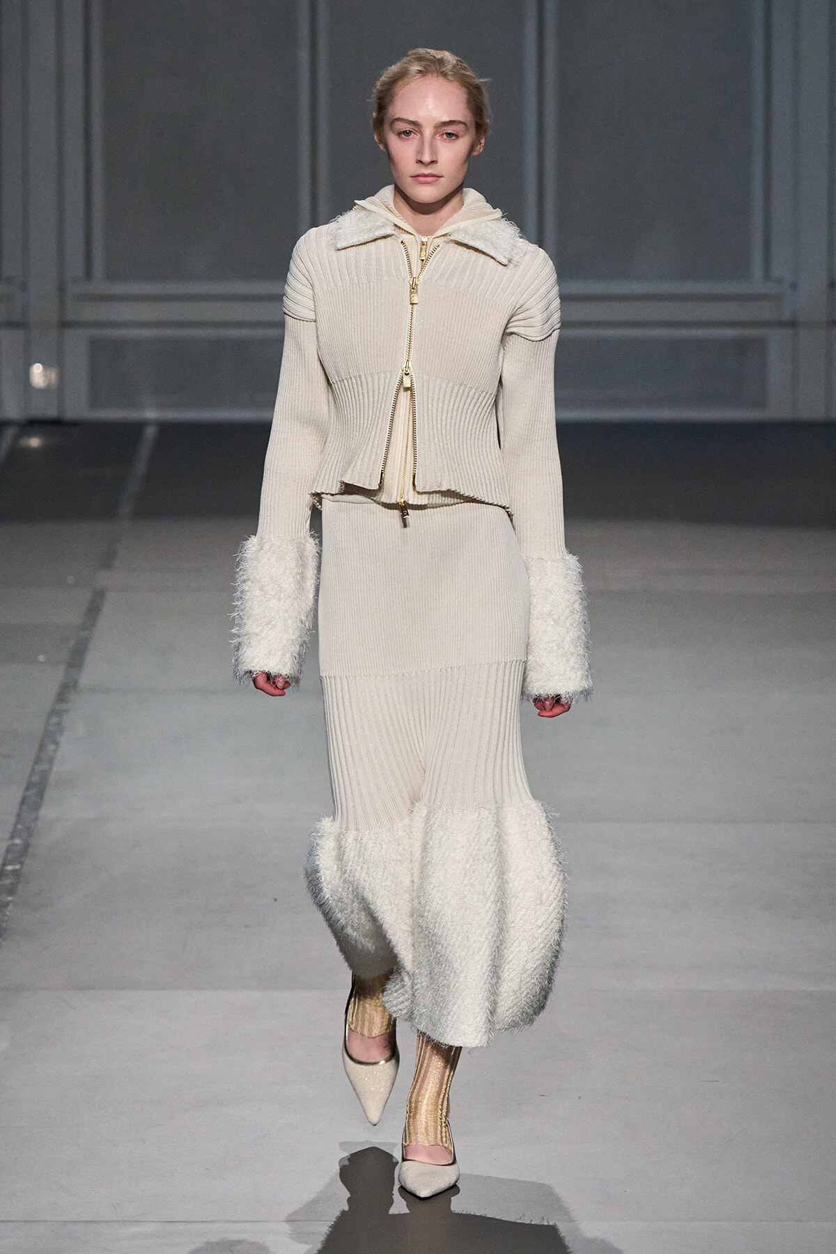 Model on a runway wearing a cream ribbed knit cropped jacket with a gold zipper, matching knit skirt, and fluffy fur cuffs; beige shoes.