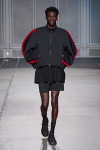 Male model walks the runway wearing an oversized black jacket with red piping, black shorts, and black sneakers with tall socks, in a minimalist gray backdrop.