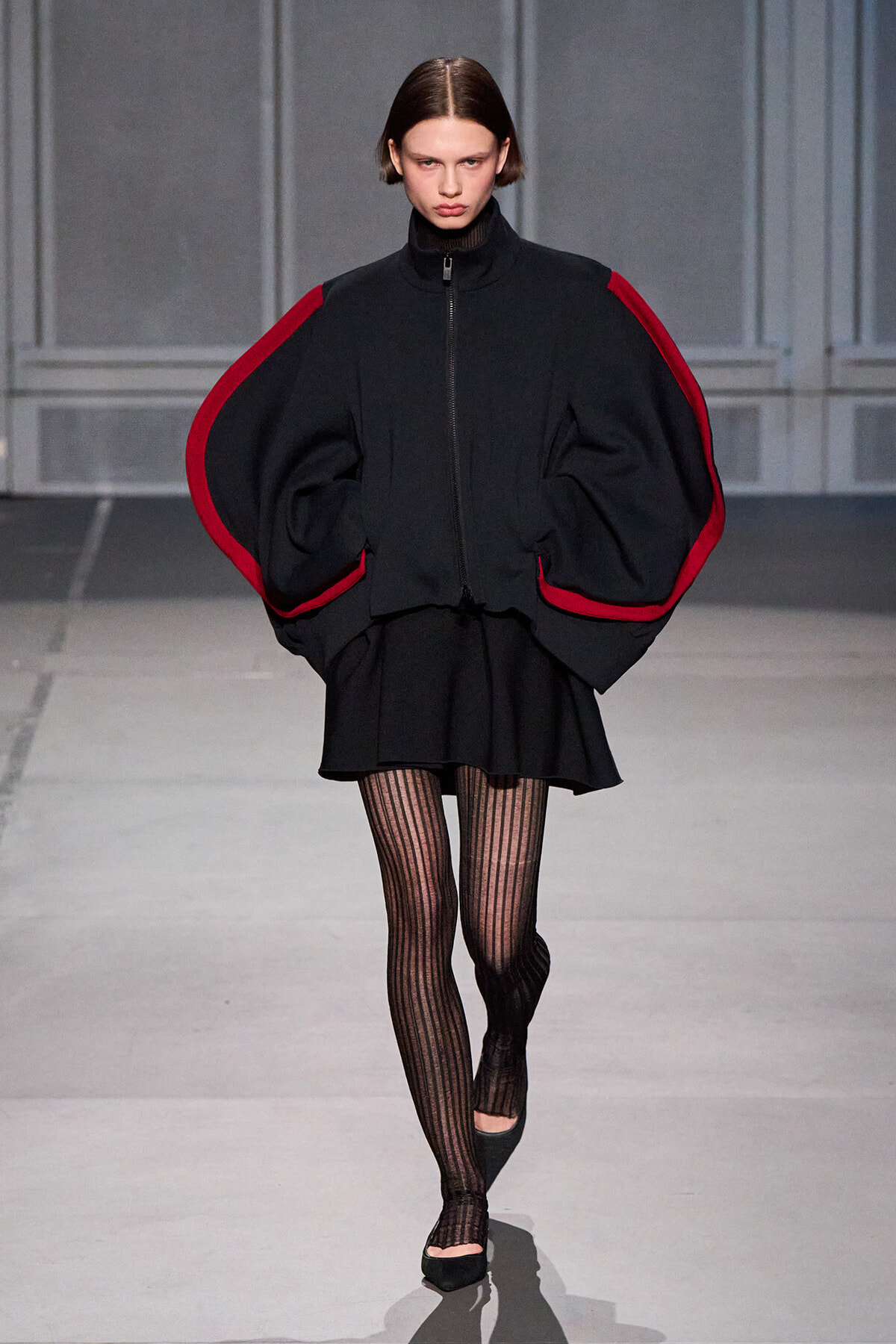 Model walking a runway in a black oversized cape with red trim, paired with a short black skirt and striped tights.