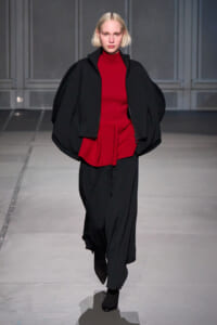 Model on a runway wearing a red knit top with an oversized black cape and wide black pants.