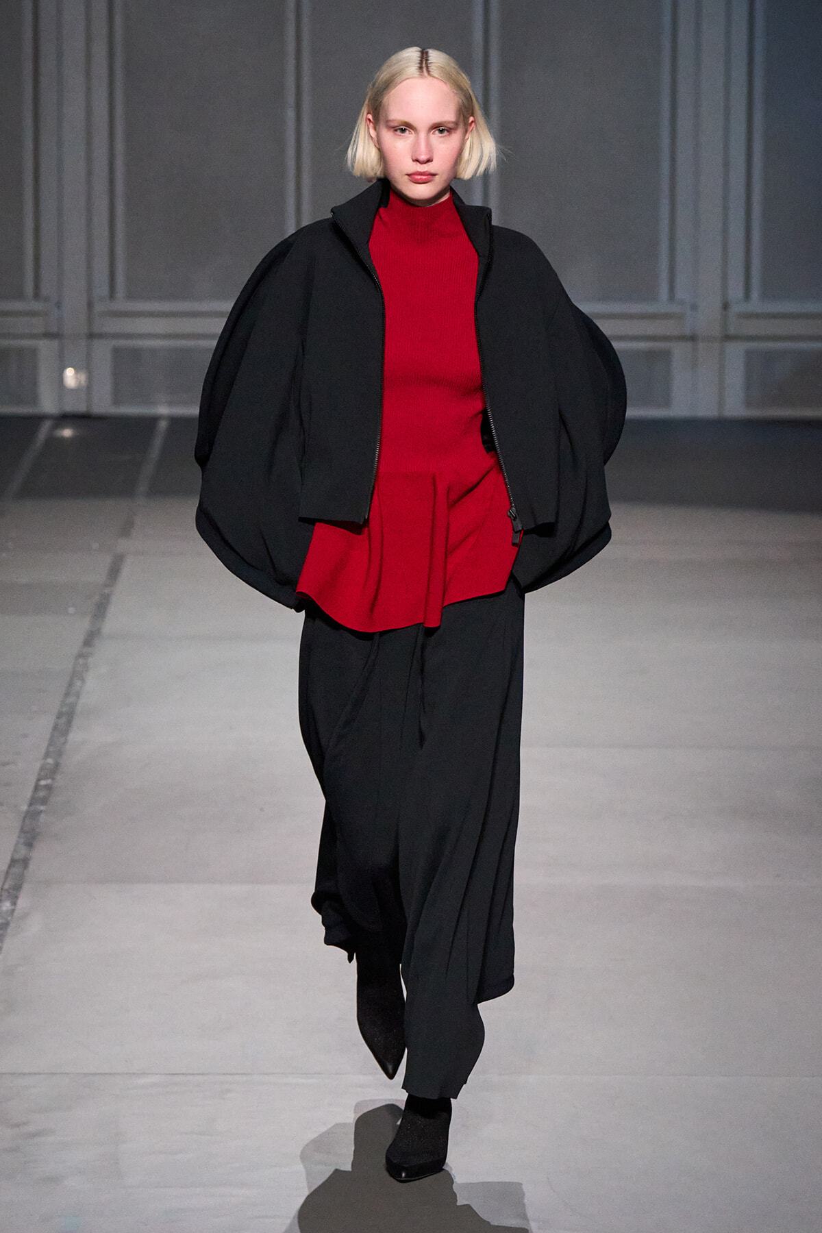 Model on a runway wearing a red knit top with an oversized black cape and wide black pants.