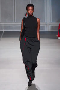 Model walking a runway in a sleeveless black knit top and wide black pants with red piping, carrying a black shoulder bag and wearing red fringe earrings.
