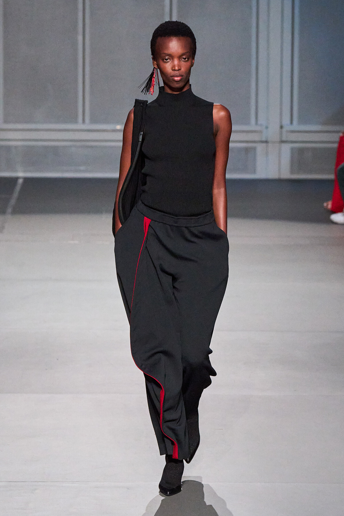 Model walking a runway in a sleeveless black knit top and wide black pants with red piping, carrying a black shoulder bag and wearing red fringe earrings.