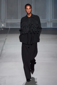 Model walks a fashion runway in an oversized black top with layered fringe, paired with wide black trousers and rounded glasses.