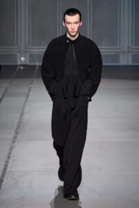 Male model walking the runway in an all-black outfit: oversized textured jacket, black top, and wide-leg pants with hands in pockets.