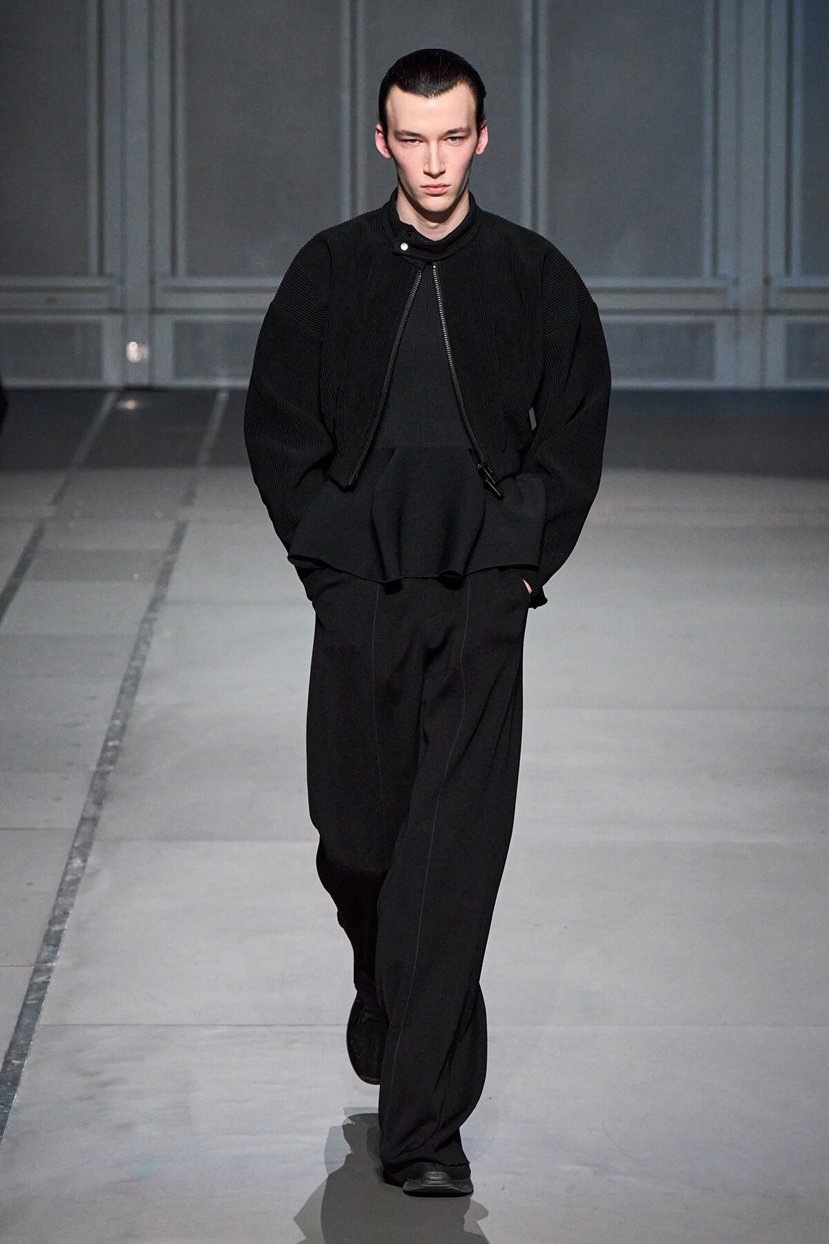 Male model walking the runway in an all-black outfit: oversized textured jacket, black top, and wide-leg pants with hands in pockets.